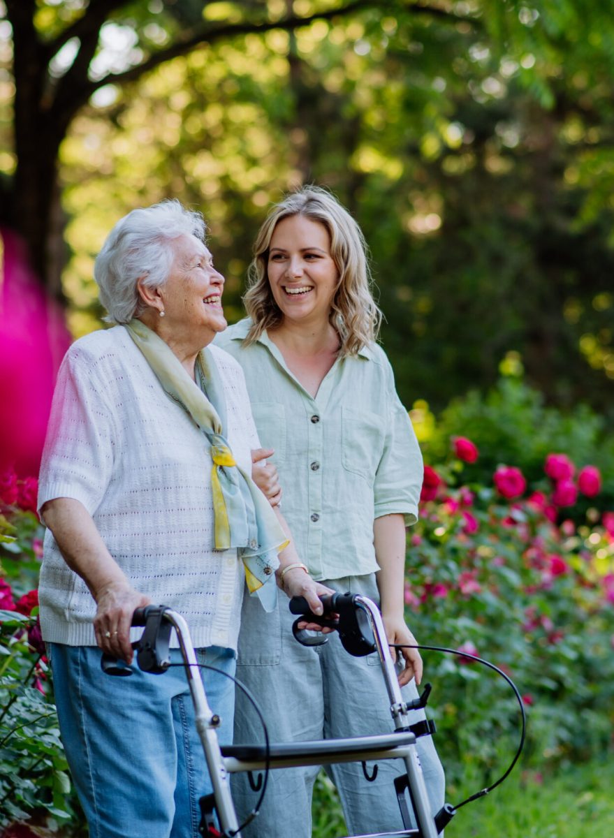 An adult granddaguhter supporting her senior grandmother when taking her for walk with walker in park in summer.